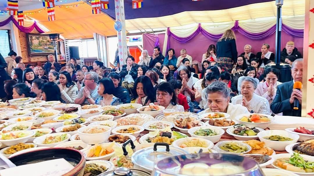 A hall full of people praying behind long tables of food offerings